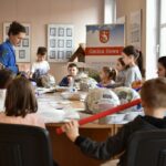 Grupa dzieci zgromadzona wokół stołu podczas wykonywania prac plastycznych. Obok stoi instruktorka