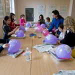 Zajęcia plastyczne. Dzieci siedzą przy stole. Każde z nich ma balon. Obok stoi instruktorka